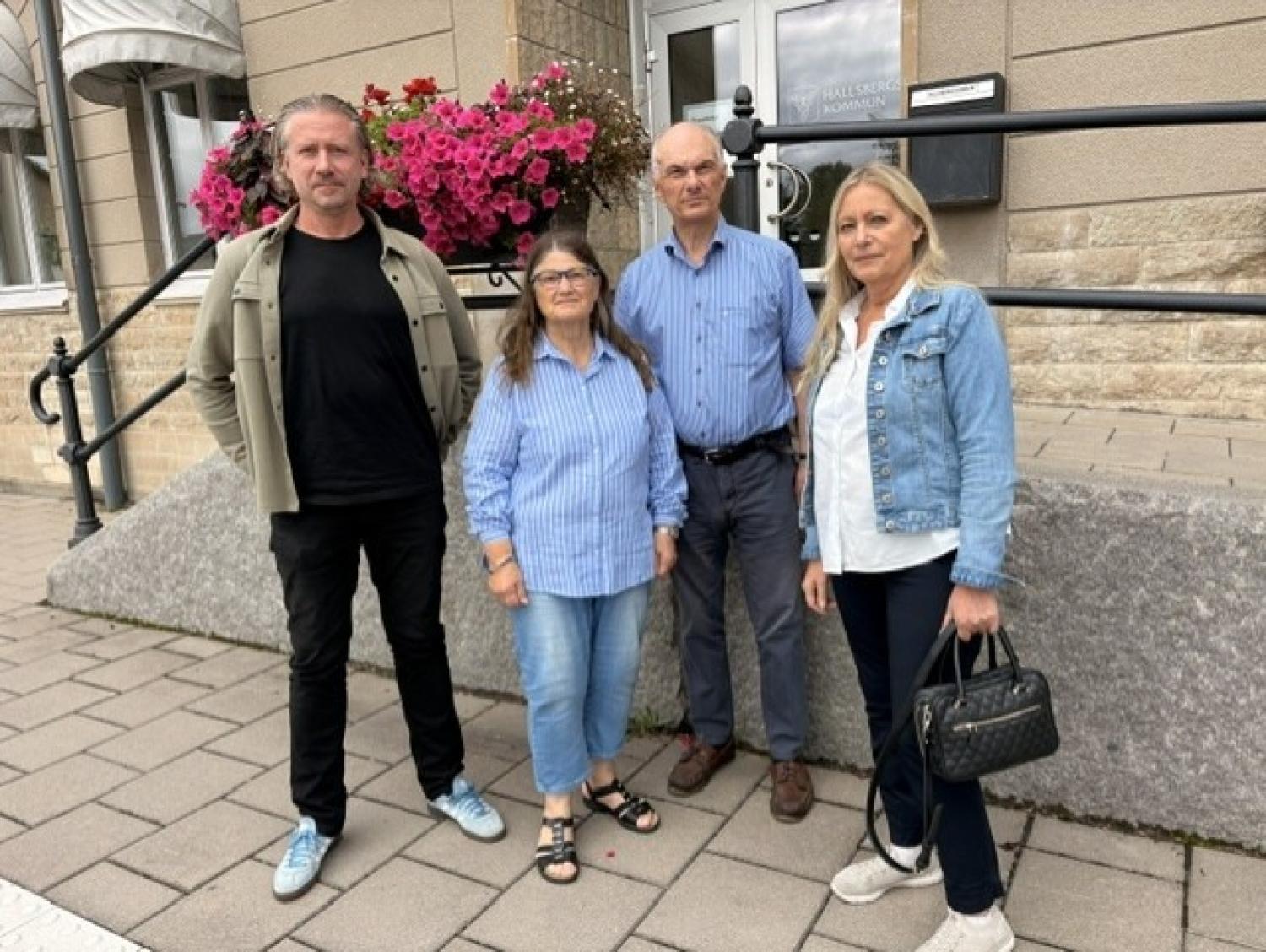 Fyra vuxna står utanför en byggnad med en blomlåda, klädda i vardagskläder, och tittar in i kameran.