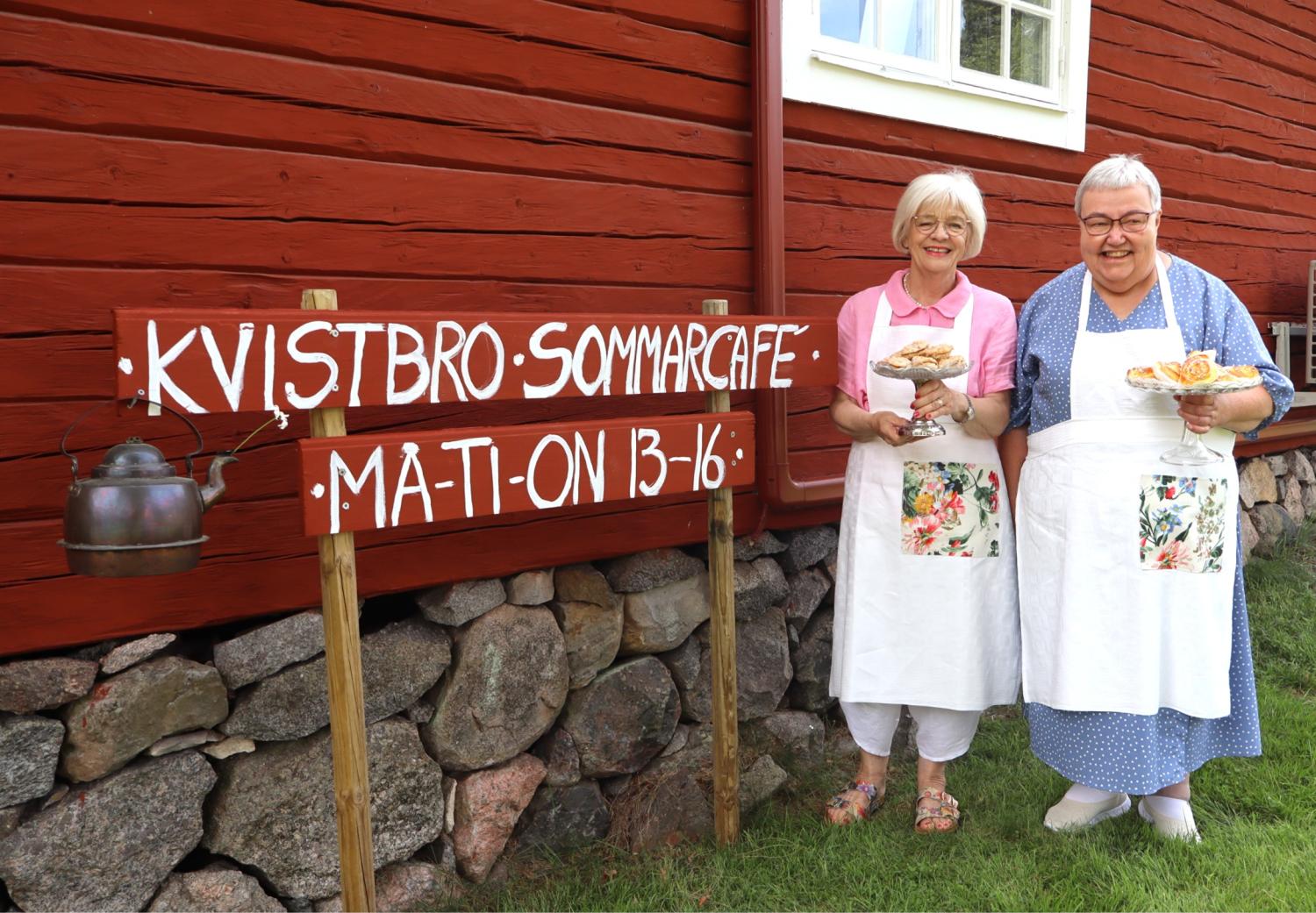 Två kvinnor i förkläden står vid en röd träbyggnad och håller i tallrikar med bakverk bredvid en skylt där det står ”KVISTBRO SOMMARCAFE MAT-ON 13–16”, med en vattenkokare hängande på skylten.