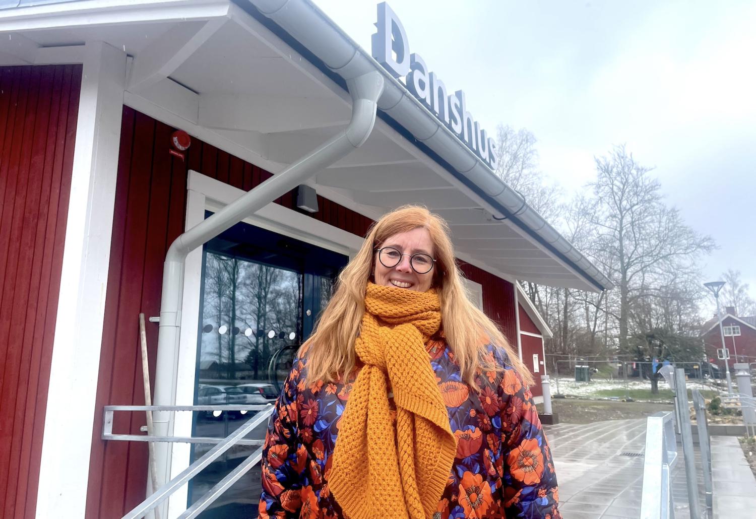 En kvinna med glasögon och en gul halsduk ler framför en röd byggnad med en skylt där det står 