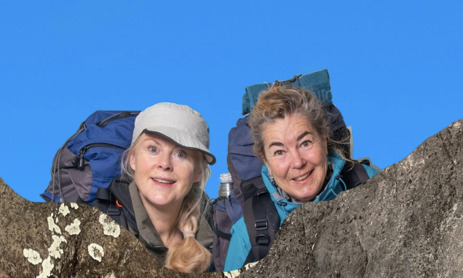 Två kvinnor med stora ryggsäckar ler och kikar över en stenig avsats mot en klarblå himmel.