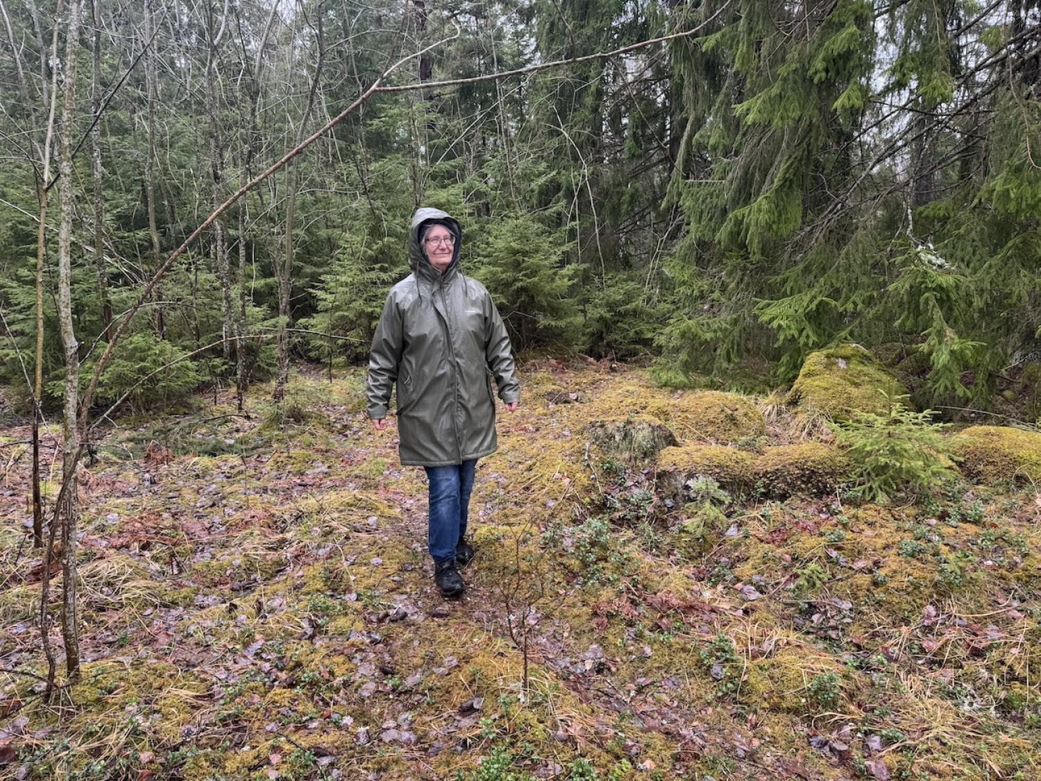 En person iförd regnrock och hatt står och ler i en frodig, grön skog med mossbeklädd mark.