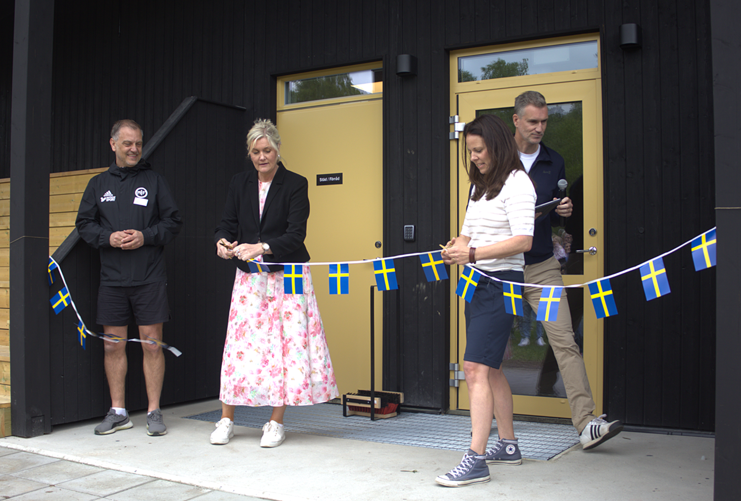 Fyra personer står utanför en byggnad och klipper ett band dekorerat med svenska flaggor under vad som ser ut att vara en invigningsceremoni.