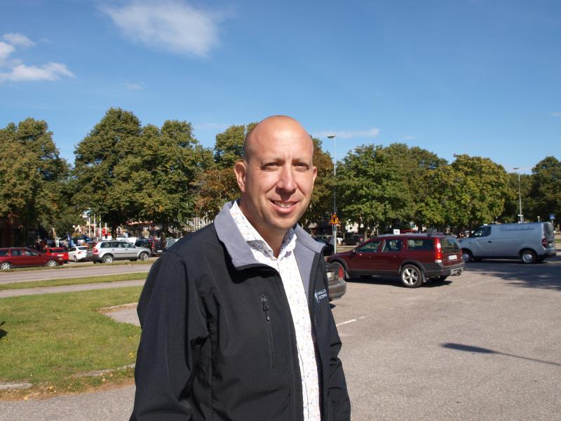 En man i svart jacka står på en parkeringsplats med bilar och träd i bakgrunden en solig dag.