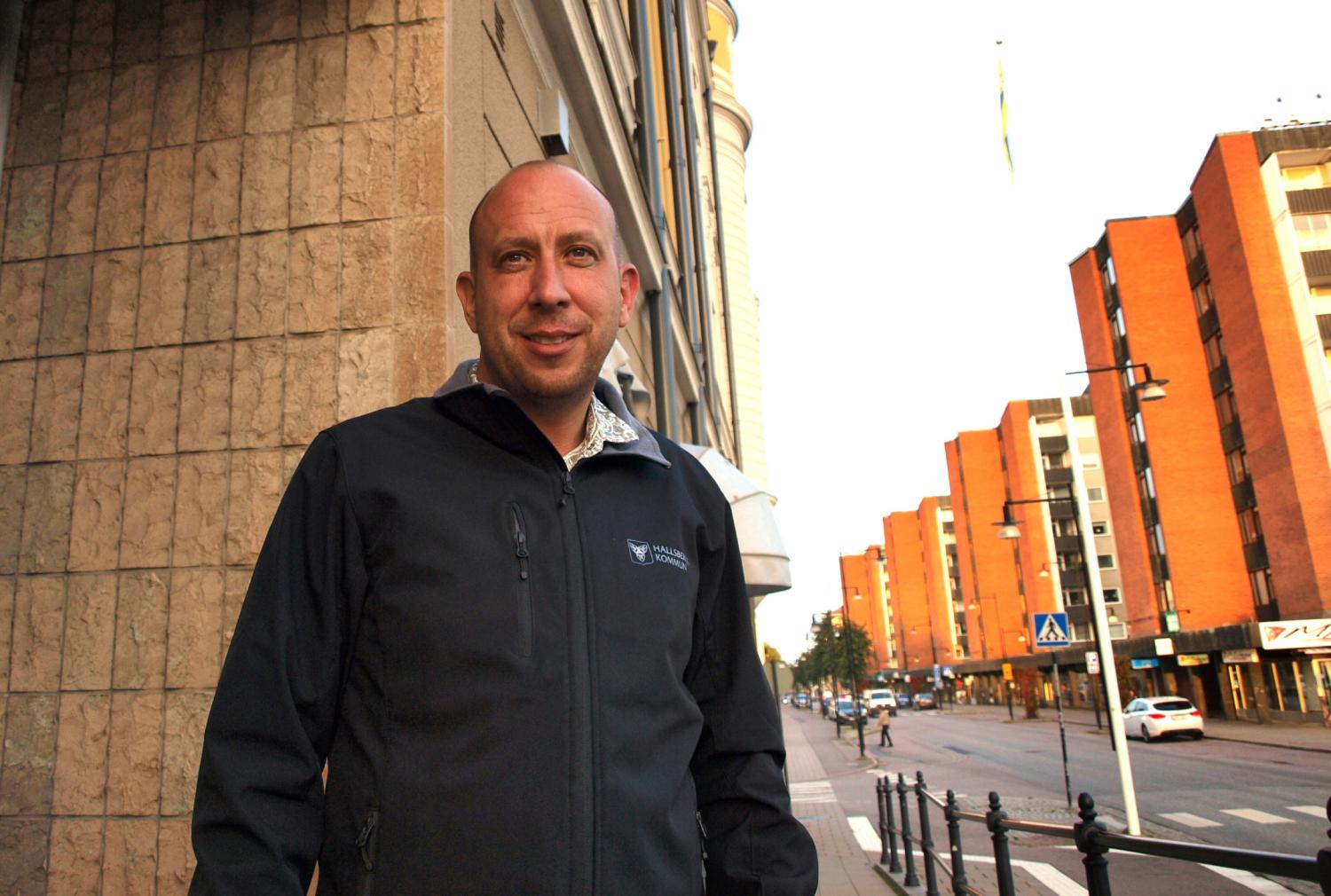 En man i mörk jacka står på en trottoar i en stad bredvid en byggnad, med orangefärgade flerfamiljshus och en gata i bakgrunden.