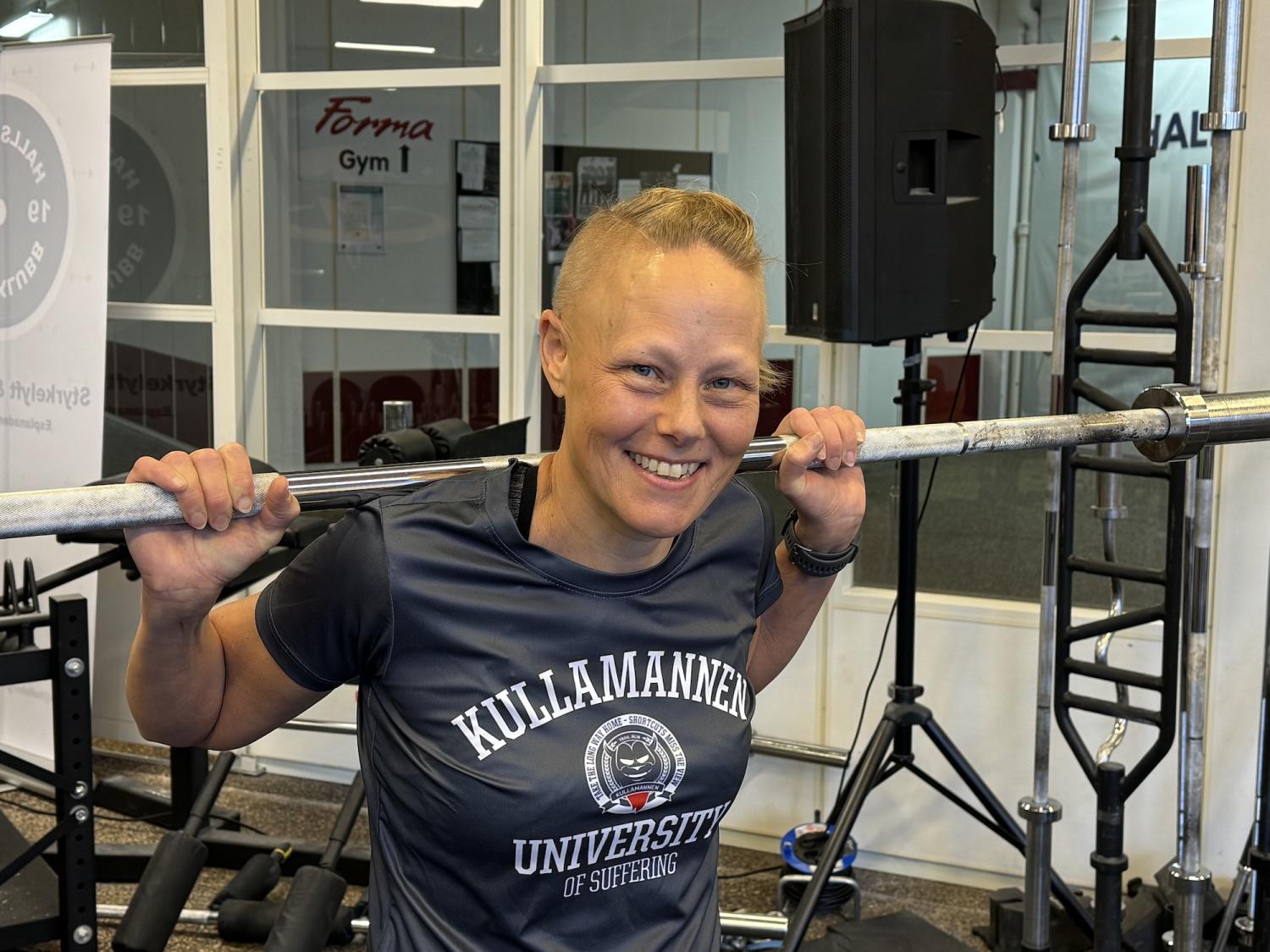 Person på ett gym som håller en skivstång på sina axlar, ler, bär en 