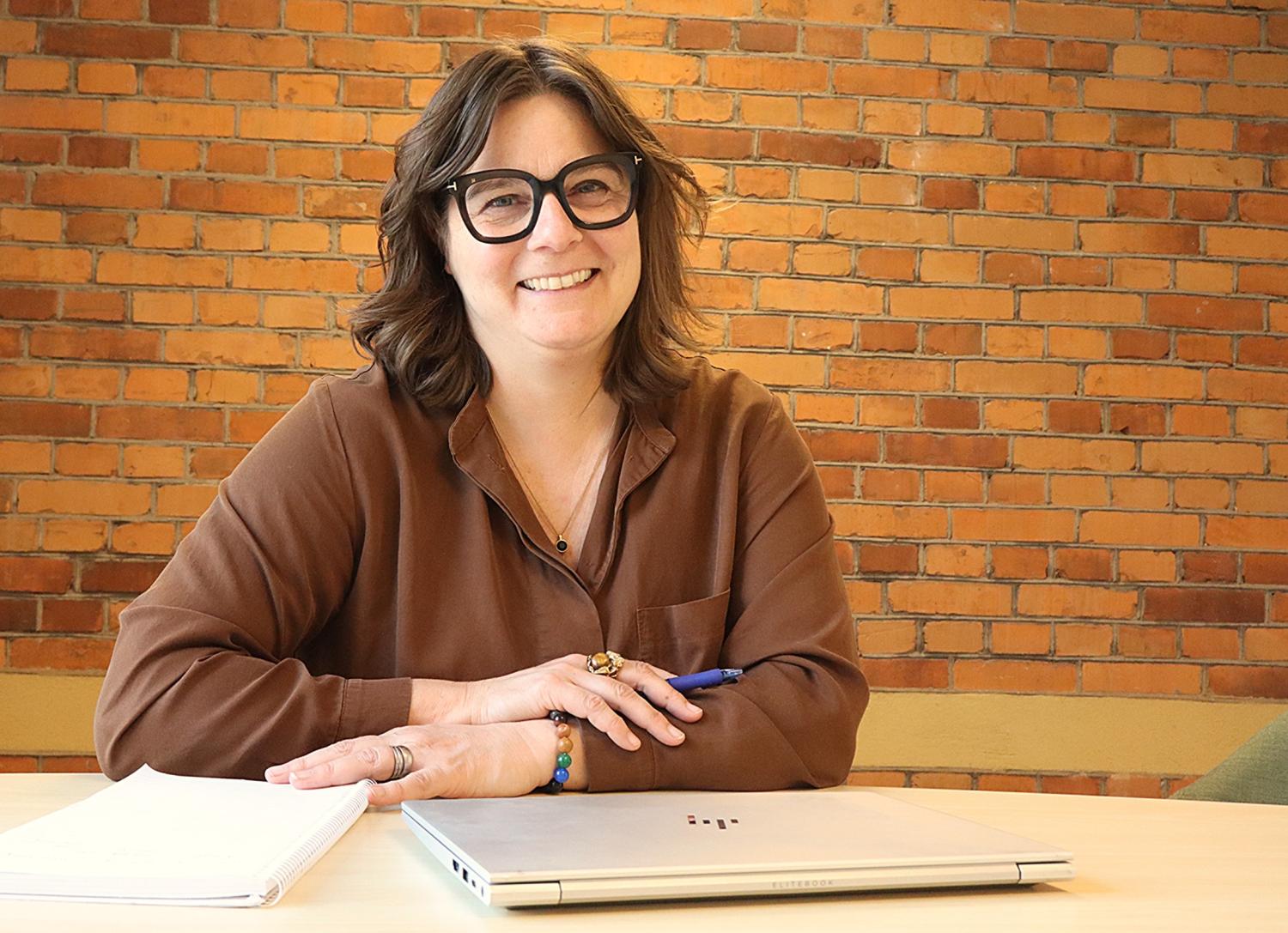 En person med glasögon sitter vid ett bord med en bärbar dator, anteckningsbok och penna och ler mot en bakgrund av en tegelvägg.