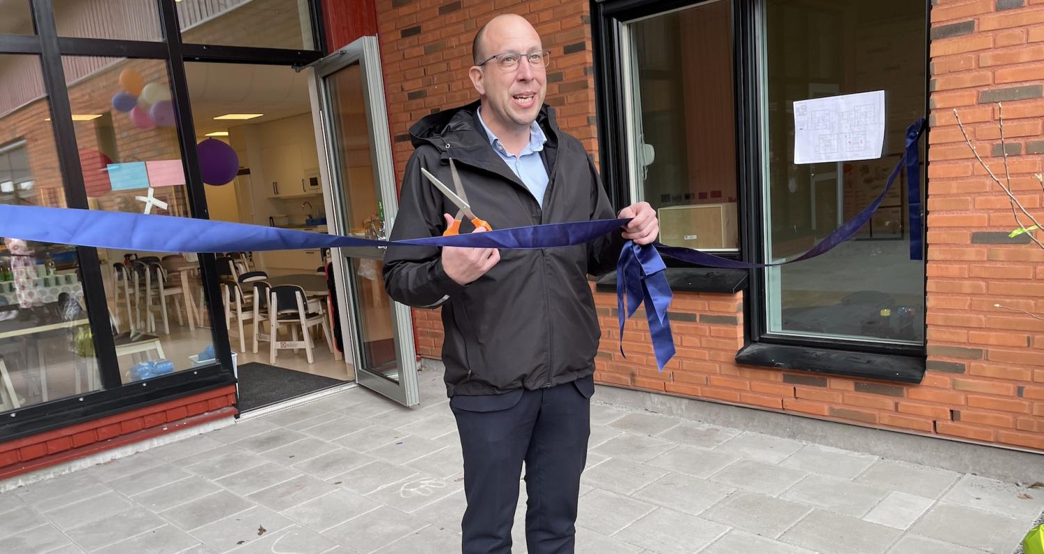 En man i glasögon och en jacka klipper ett blått band vid en byggnadsingång under en ceremoniell händelse.