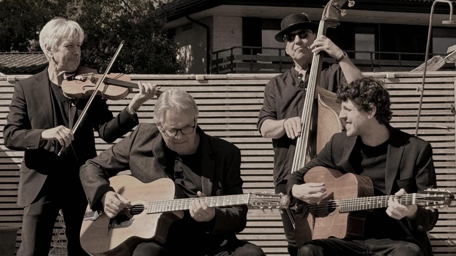 Fyra musiker uppträder utomhus och spelar violin, kontrabas och två akustiska gitarrer, alla är mörkklädda och sitter framför ett staket av träribbor.