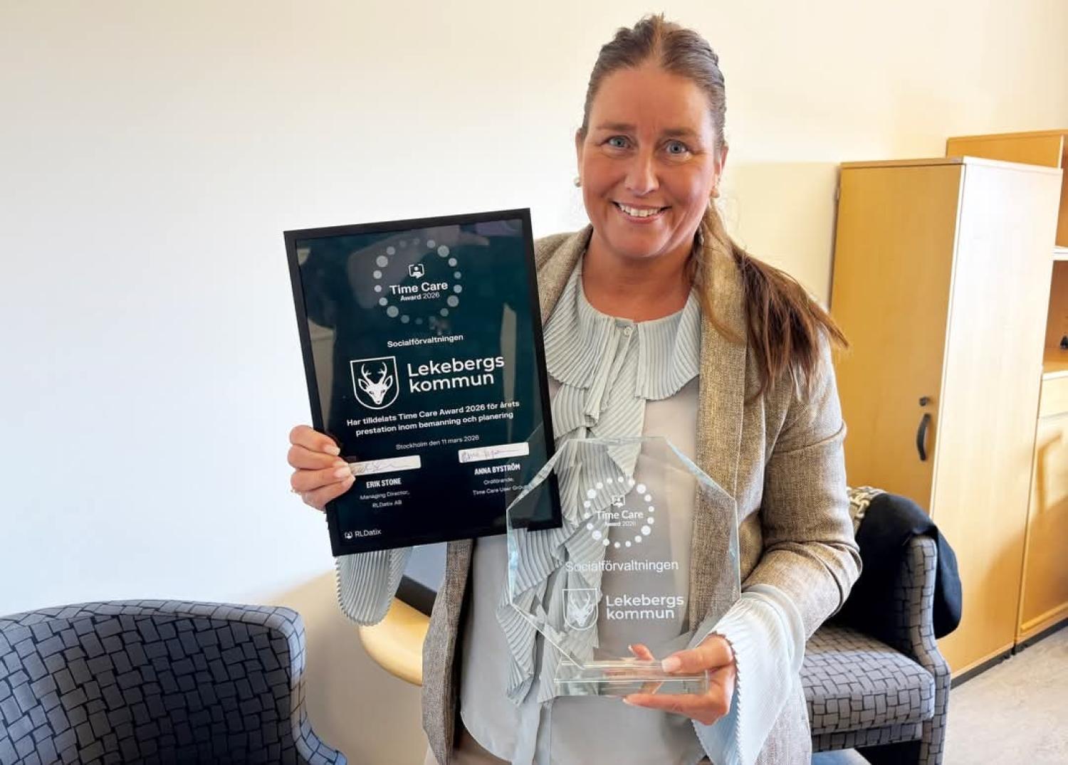 En kvinna står inomhus och ler, med ett inramat certifikat i ena handen och ett pris i klarglas i den andra, båda med texten 