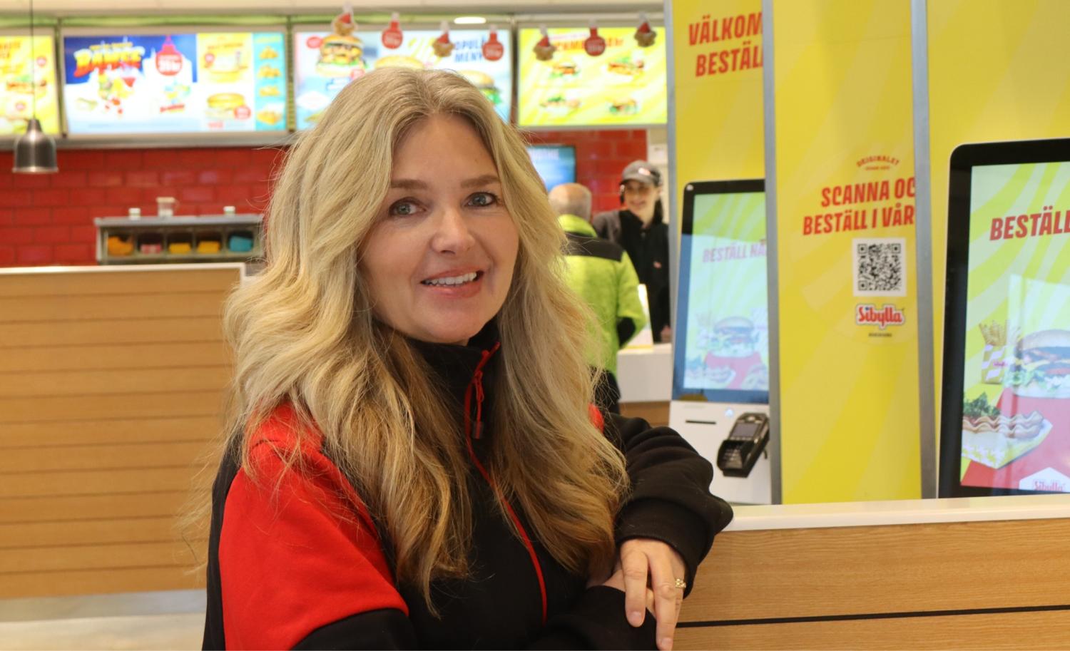 En kvinna med långt blont hår och en svart-röd jacka står inne på en snabbmatsrestaurang nära självbeställningsskärmar och menytavlor.