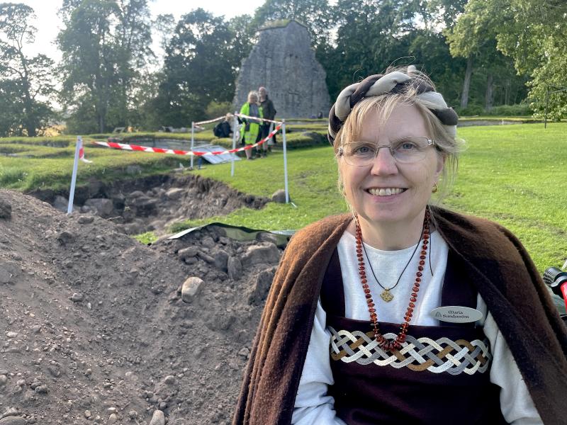 En kvinna i historisk klädsel ler nära en arkeologisk utgrävningsplats med varningstejp och ett gräsområde i bakgrunden.