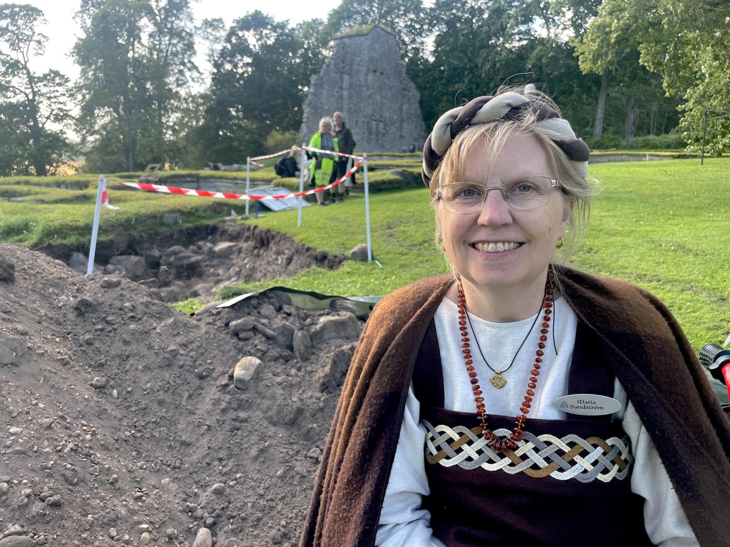 En kvinna i historisk klädsel ler nära en arkeologisk utgrävningsplats med varningstejp och ett gräsområde i bakgrunden.