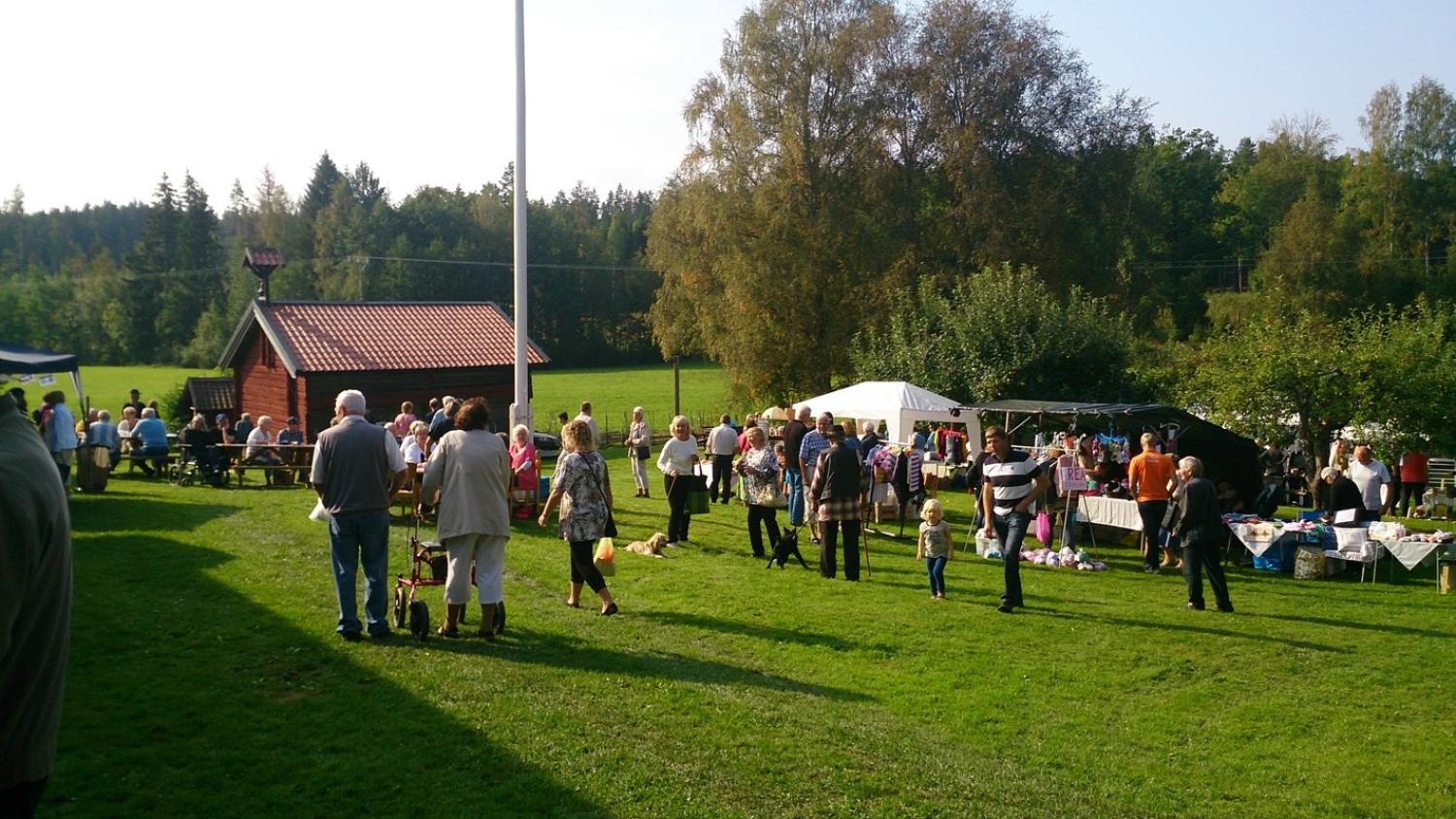 En grupp människor samlas utomhus på ett gräsfält nära flera tält och en liten byggnad, med träd och en skog i bakgrunden under en klar himmel.