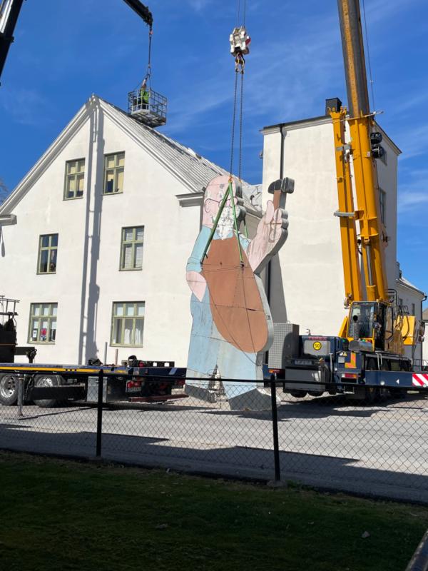 En stor väggmålning av en arbetare installeras på sidan av en vit byggnad med hjälp av kranar och en hiss under en klarblå himmel.