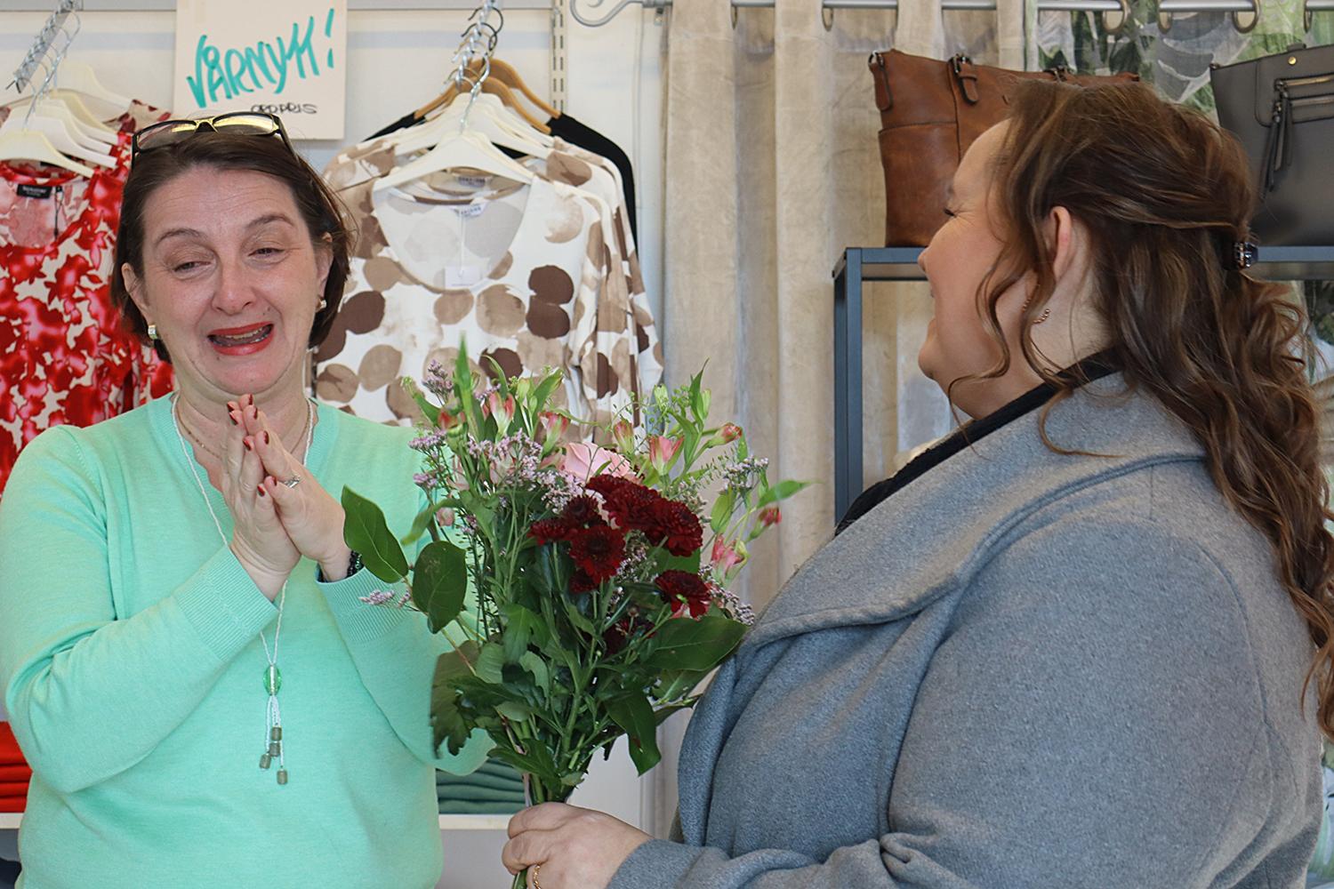 Två kvinnor står inomhus; den ena håller i en blombukett och ler, medan den andra verkar rörd med händerna knäppta nära ansiktet. Kläder och väskor syns i bakgrunden.
