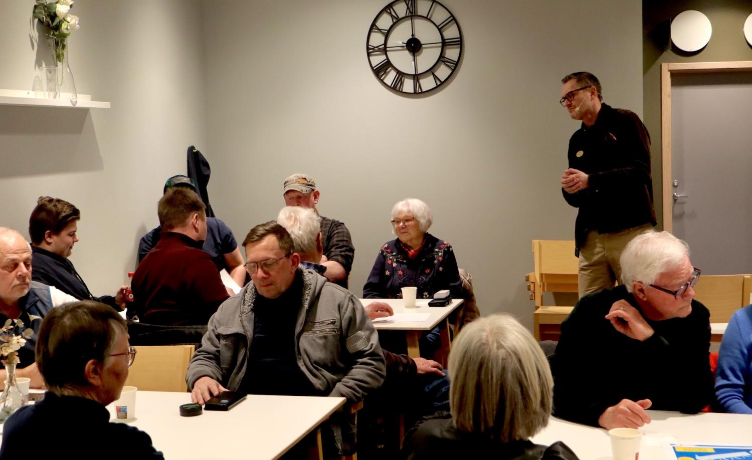 En grupp människor som sitter och står runt bord i ett rum med en väggklocka. Vissa pratar medan andra lyssnar.