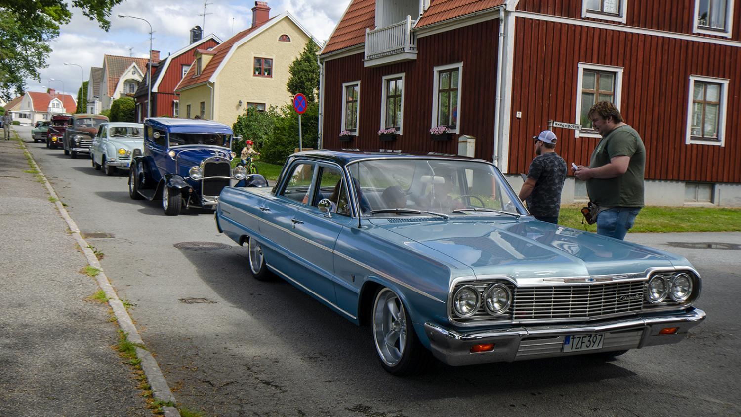 Flera veteranbilar, inklusive en ljusblå klassisk sedan, står parkerade längs en bostadsgata med människor som står i närheten.