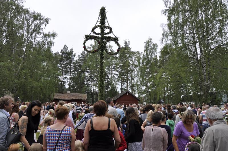 Skara människor som firar en traditionell svensk midsommarfest utomhus, med en stor majstång dekorerad med löv och blommor i bakgrunden.