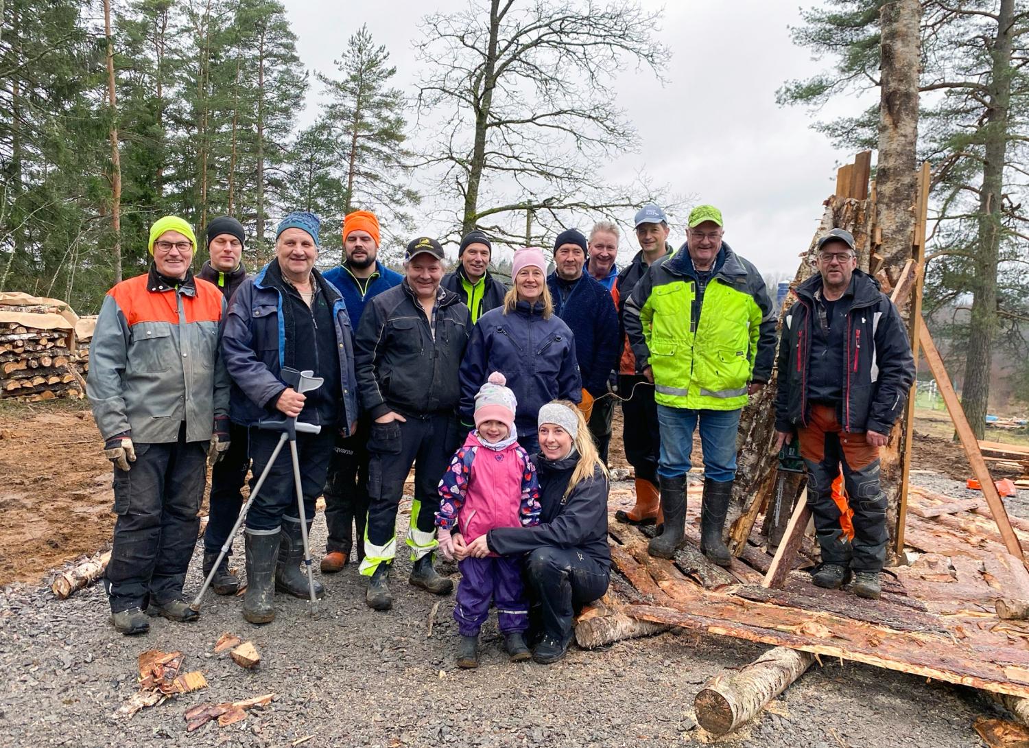 En grupp människor, däribland två barn, står utomhus i arbetskläder och skyddsutrustning vid en avverkningsplats som omges av avverkat virke och träd.