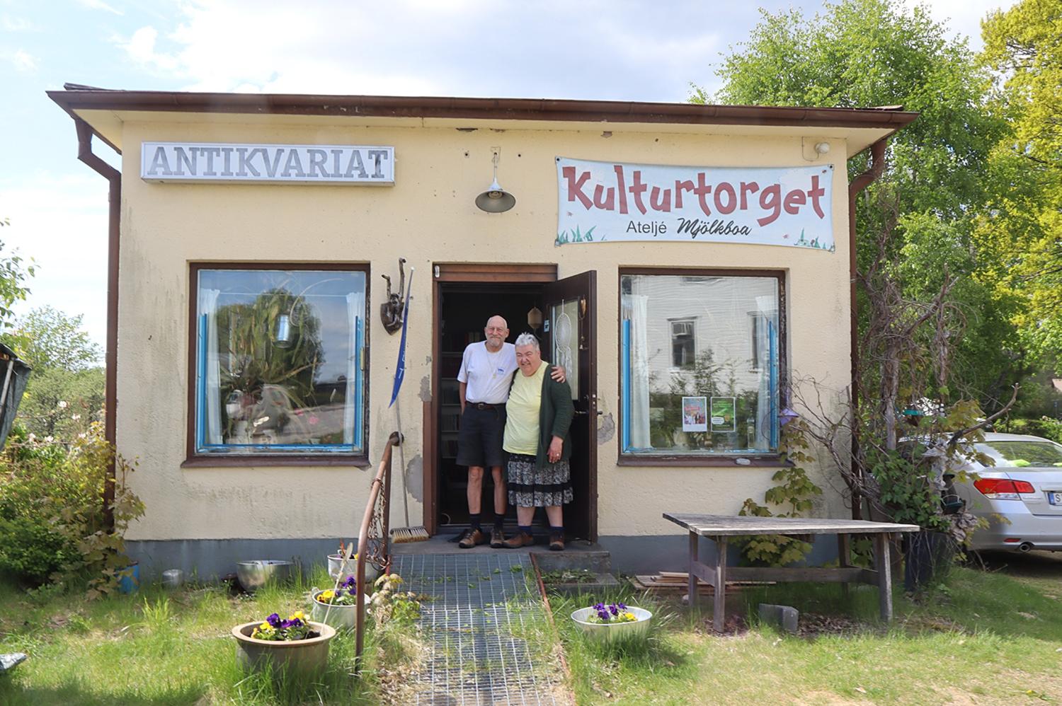Två personer står i dörröppningen till en liten byggnad med skyltar med texten 