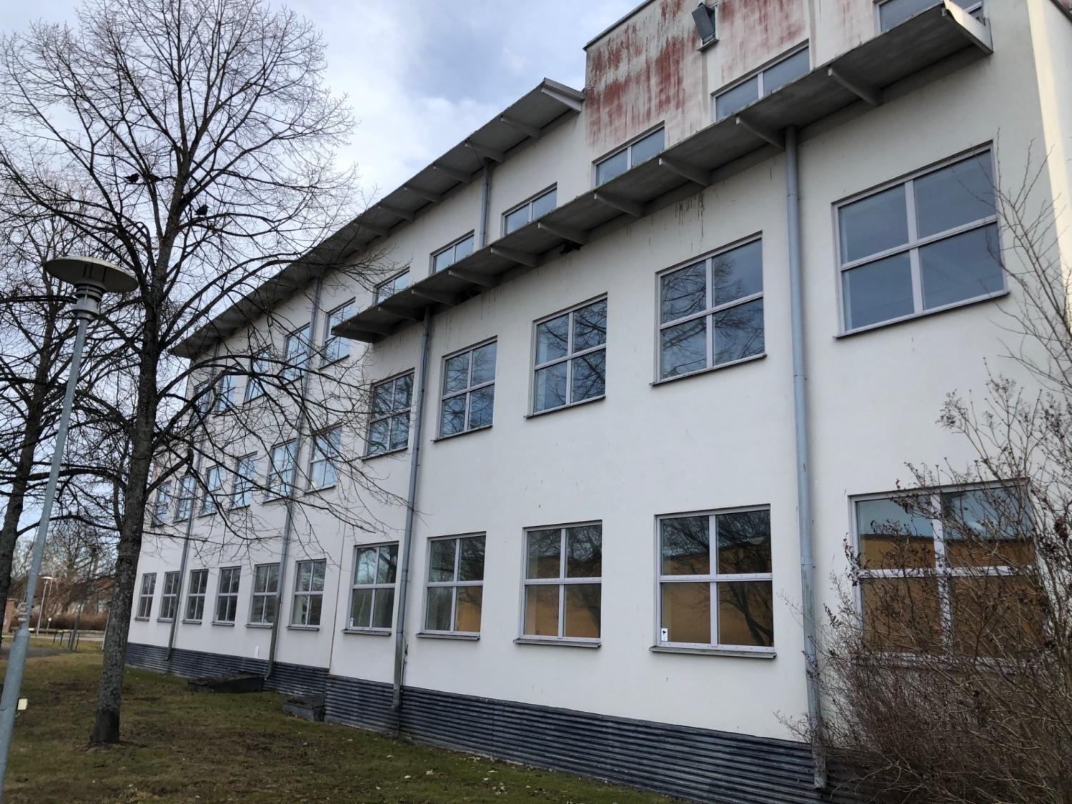 En vit byggnad i tre våningar med flera fönster, ett platt tak och synlig väderbiten skyltning på den övre fasaden; lövlösa träd och gräs i förgrunden.