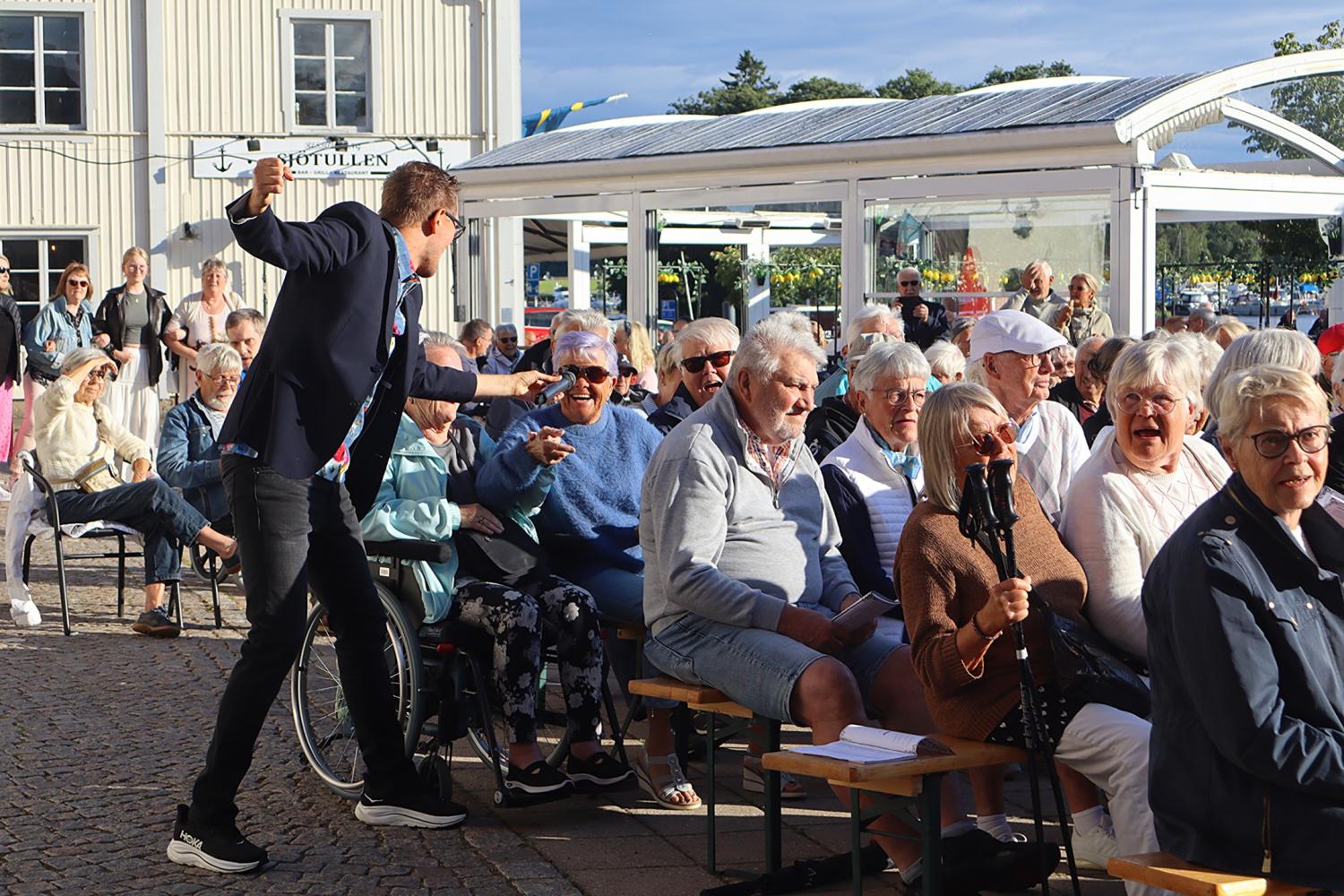 En man står upp och gestikulerar livligt mot en sittande publik bestående främst av äldre vuxna vid ett utomhusevenemang en solig dag.