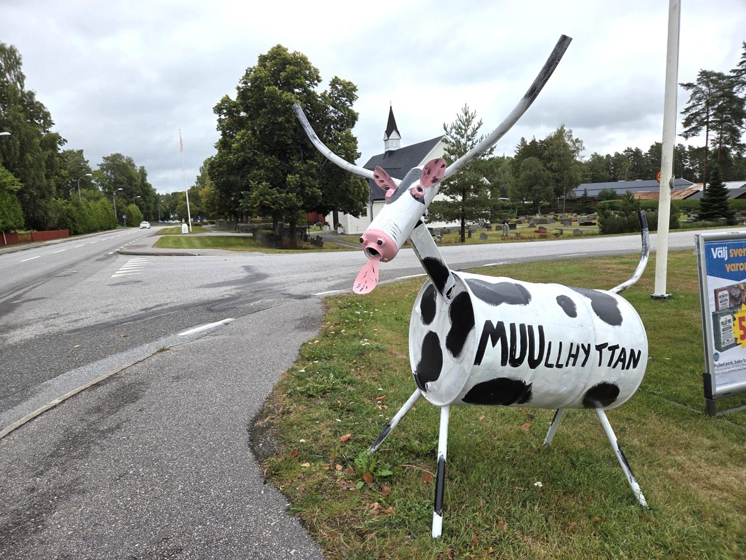 En koskulptur gjord av målade tunnor står på gräset vid en väg där det står ”MUULIHYTAN” på sidan; i bakgrunden syns träd och en kyrka.