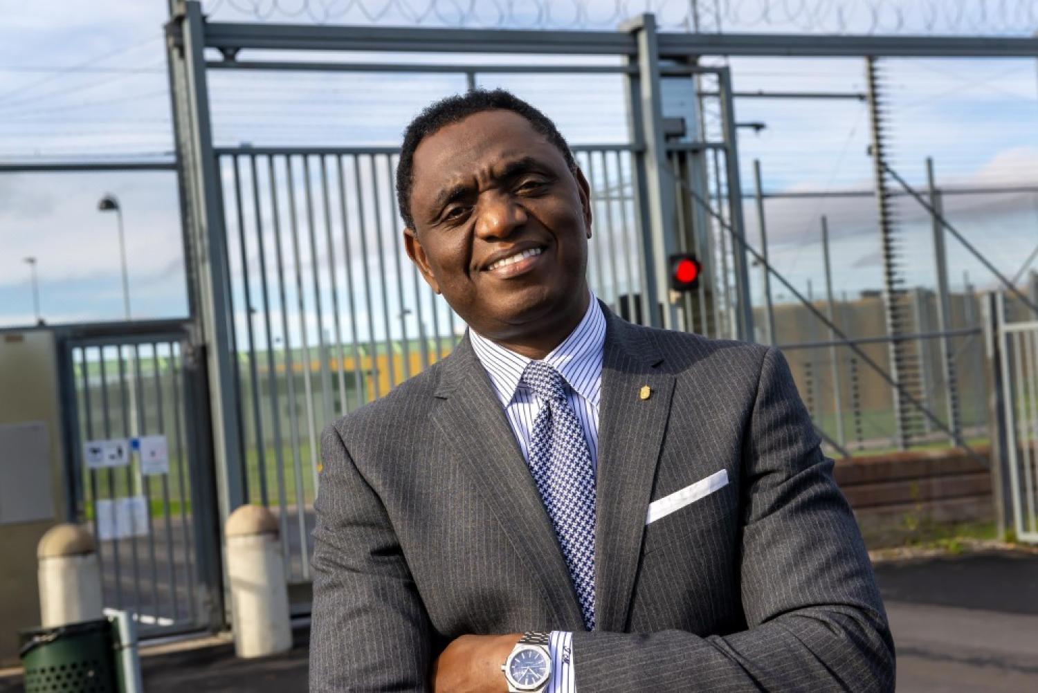 En man i kostym står med armarna i kors framför en inhägnad grind och taggtråd.