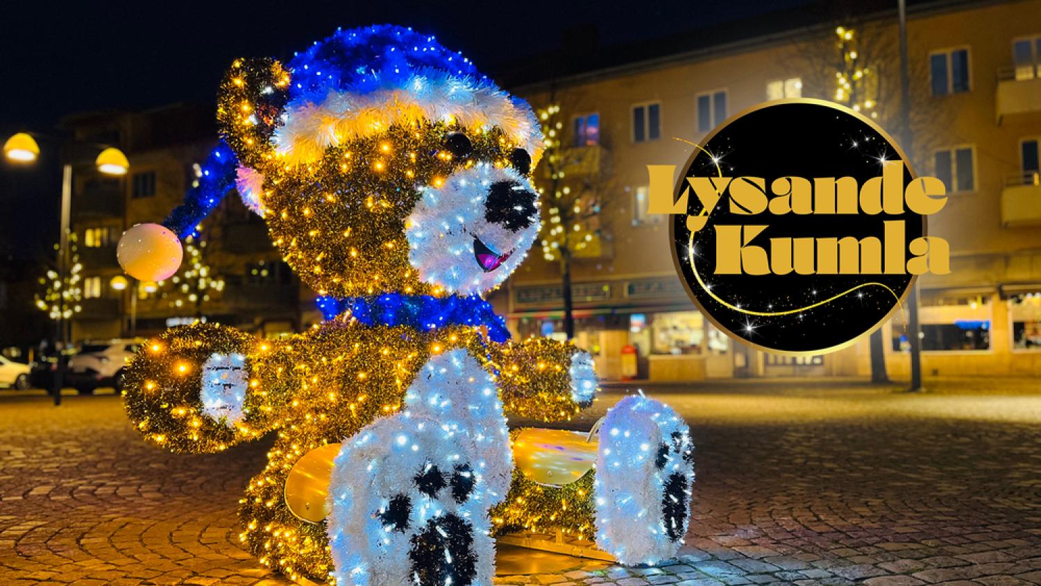 En björnformad semesterljusskärm med blå hatt sitter på en kullerstensgata på natten. Närliggande byggnader är mjukt upplysta. På en skylt står det 