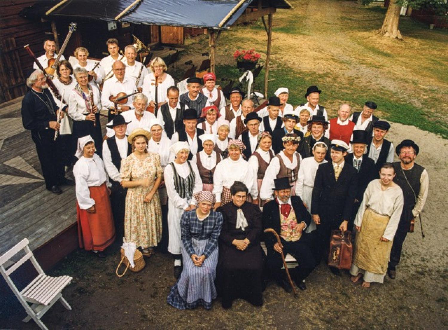 En grupp människor klädda i traditionella folkdräkter poserar utomhus, några står på trätrappor, med träd och en byggnad i bakgrunden.