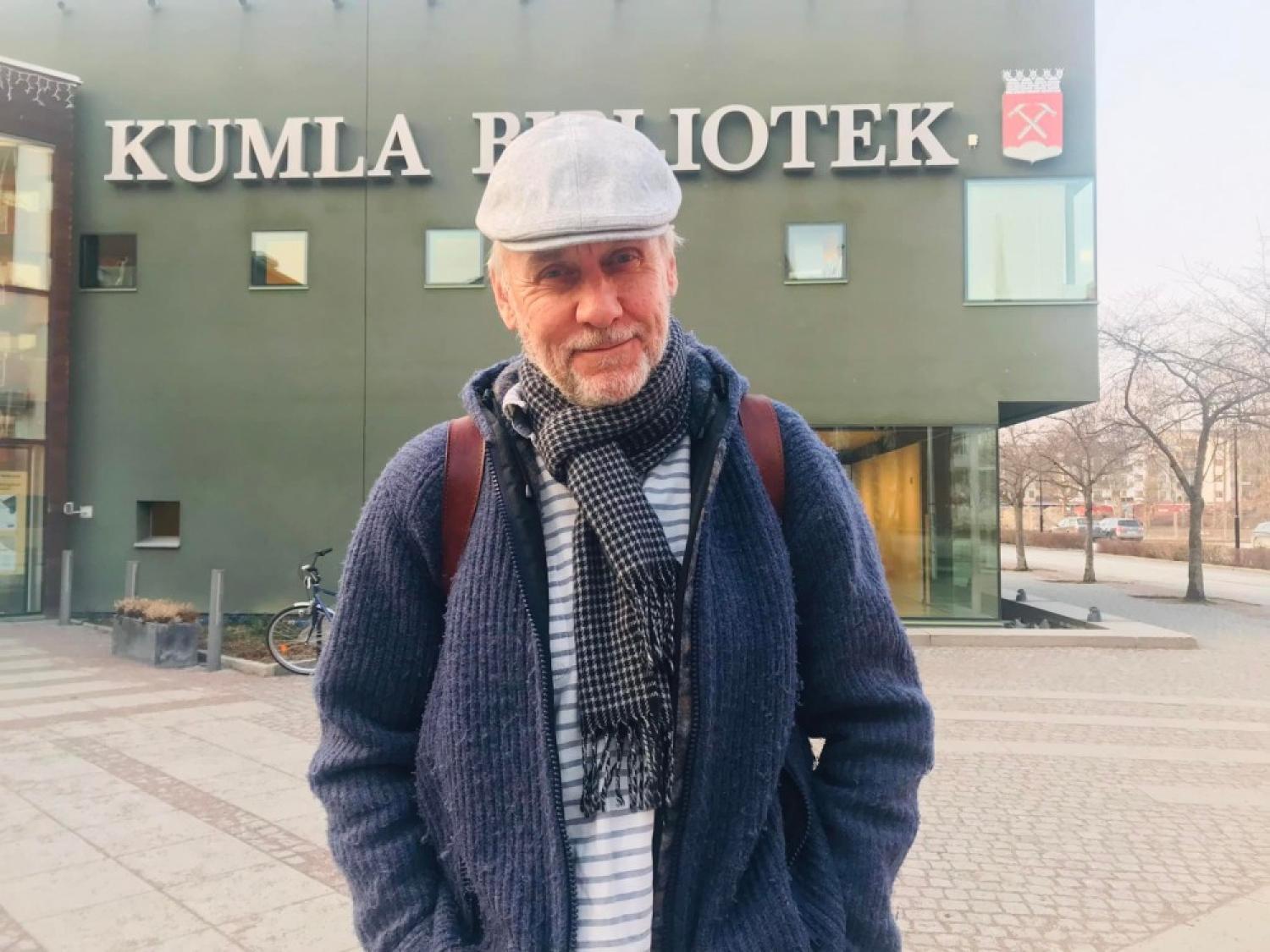 En man står utanför Kumla Bibliotek iförd en grå keps, en blå tröja och en halsduk. Bakgrunden visar biblioteksbyggnaden och ett tomt lusthusområde.