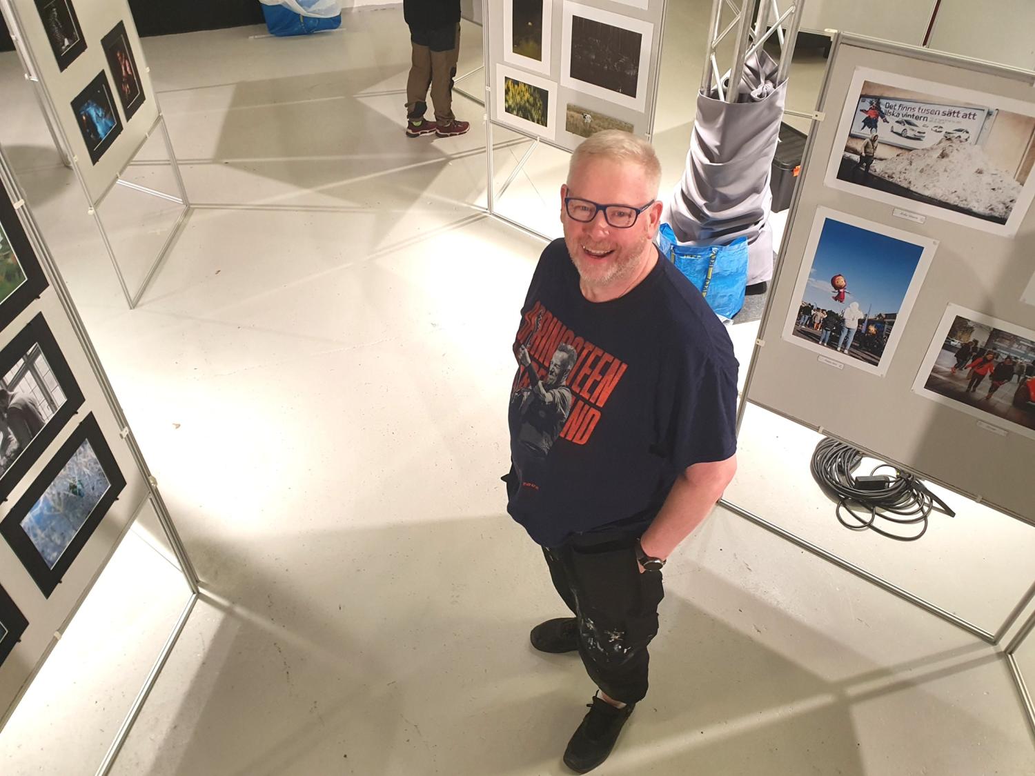 En man med glasögon och skägg står och ler i ett konstgalleri, omgiven av fotoutställningar på vita paneler.