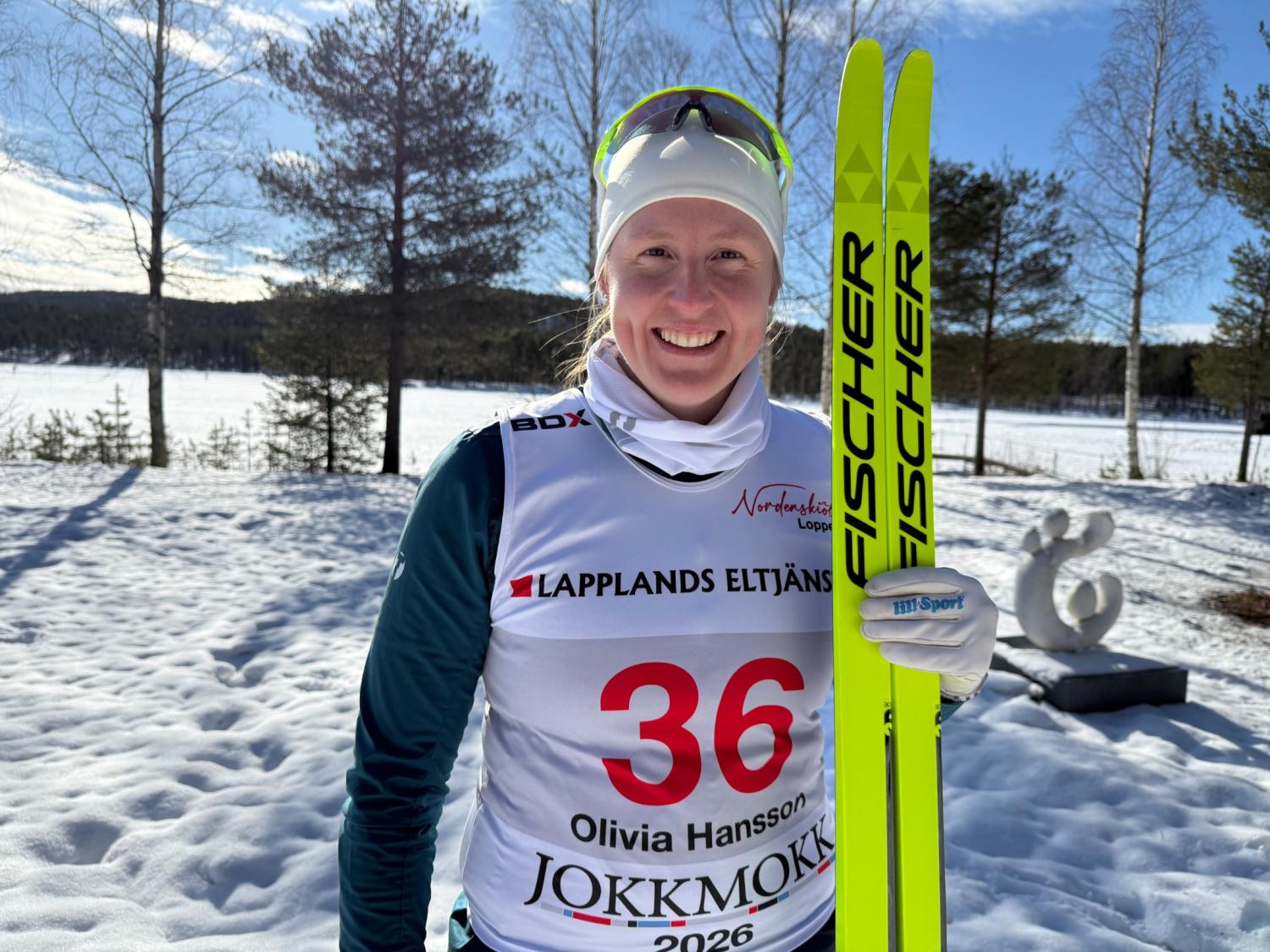 En längdskidåkare med numrerad startnummerlapp och vinterkläder står utomhus på snö med gula skidor i handen, med träd och en klar himmel i bakgrunden.