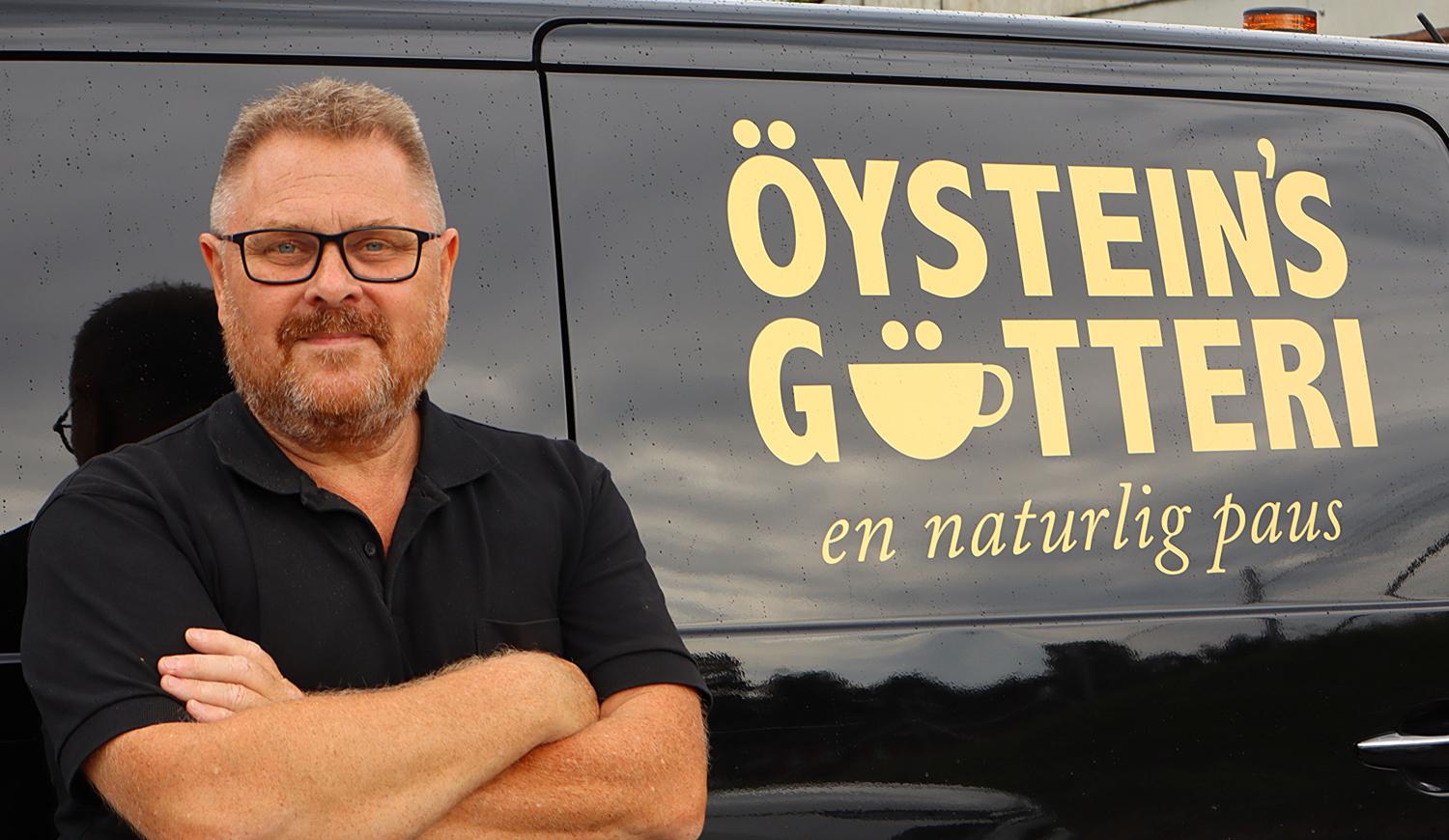 En man med glasögon och skägg står framför en svart skåpbil med texten 