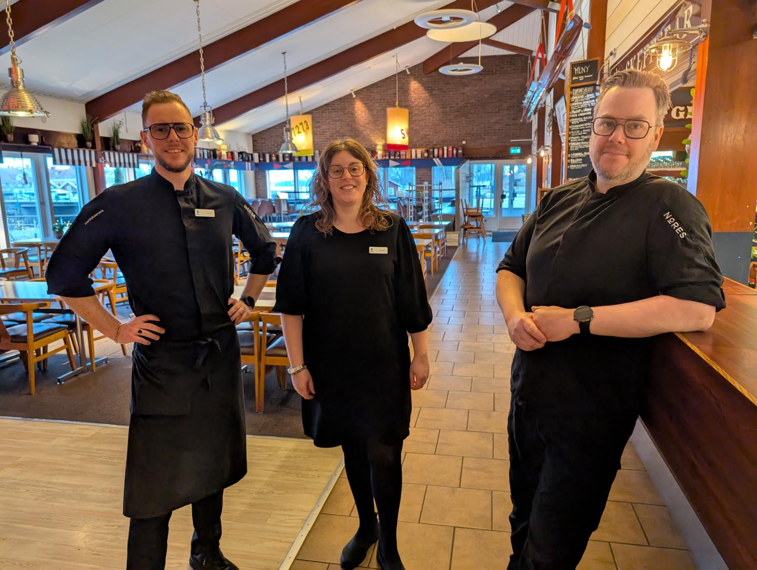 Tre restauranganställda i svarta uniformer står och ler i en rymlig, väl upplyst matsal med bord och stolar i trä.