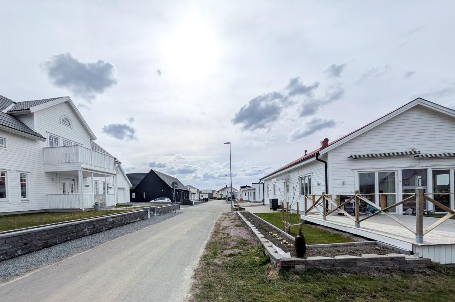 En förortsgata kantad av moderna vita hus under en molnig himmel, med en asfalterad väg och några gräsmattor i blickfånget.