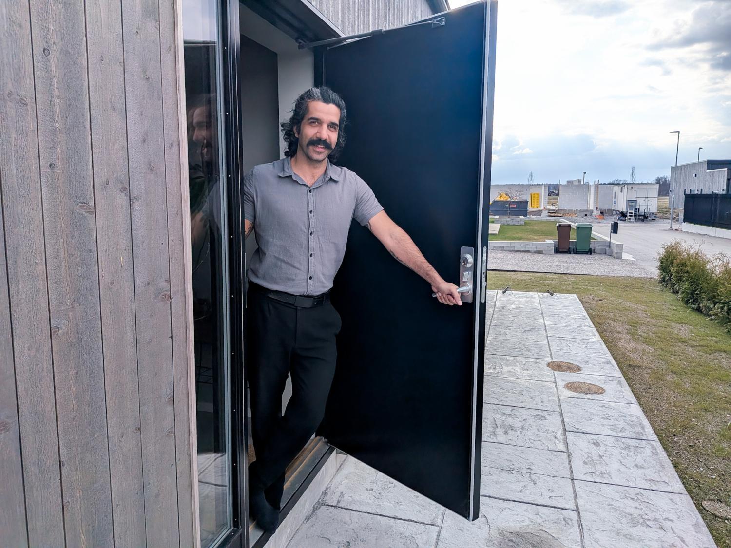 En man med mustasch står i en dörröppning, håller dörren öppen och tittar ut. Miljön ser ut att vara en modern byggnad med en gångväg utomhus och en mulen himmel.