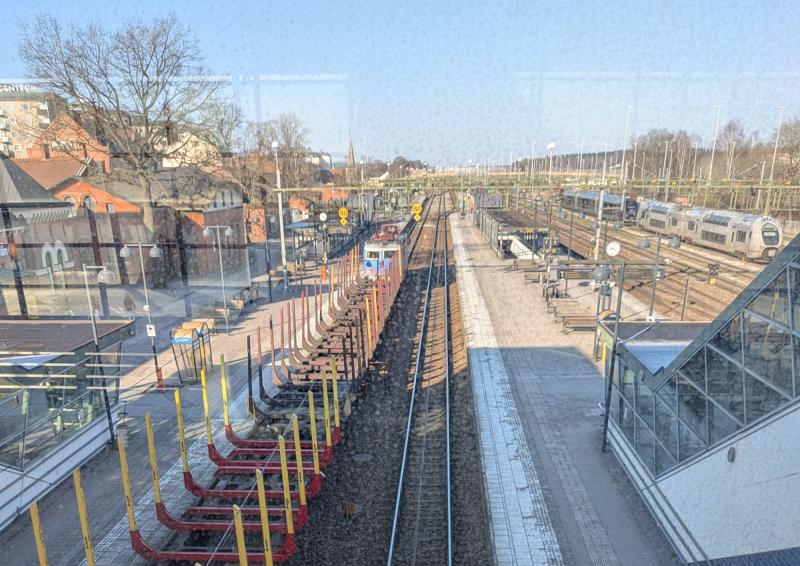 Vy över en tågstation med tomma spår, godstågsvagnar, några passagerare på perronger och byggnader i bakgrunden under en klar himmel.
