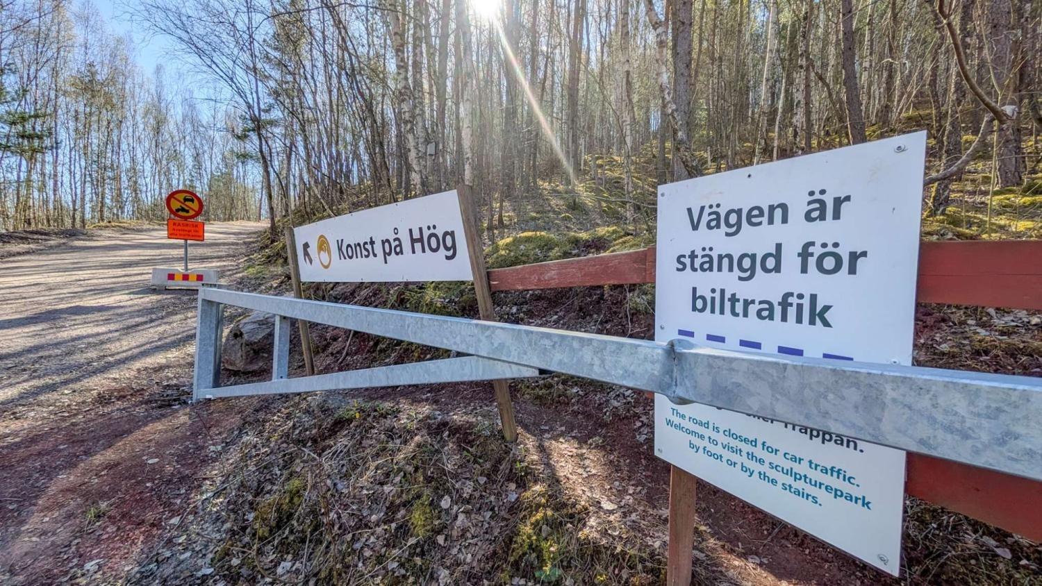 En skogsstig med skyltar på svenska, varav en anger att vägen är avstängd för biltrafik och en annan visar ”Konst på Hög”. Träd och en avspärrning syns.