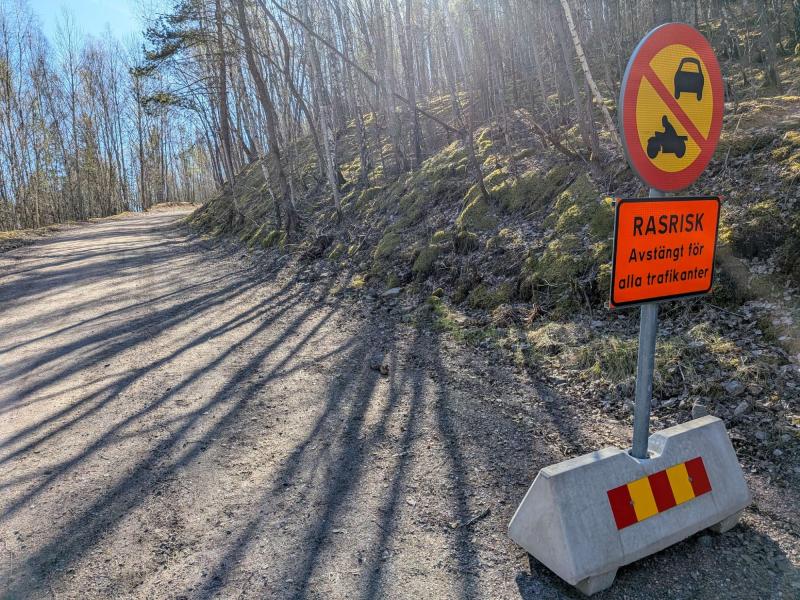 En grusväg i en skog är avstängd för bilar och motorcyklar, med en varningsskylt på svenska om skredrisk och en betongbarriär som blockerar vägen.