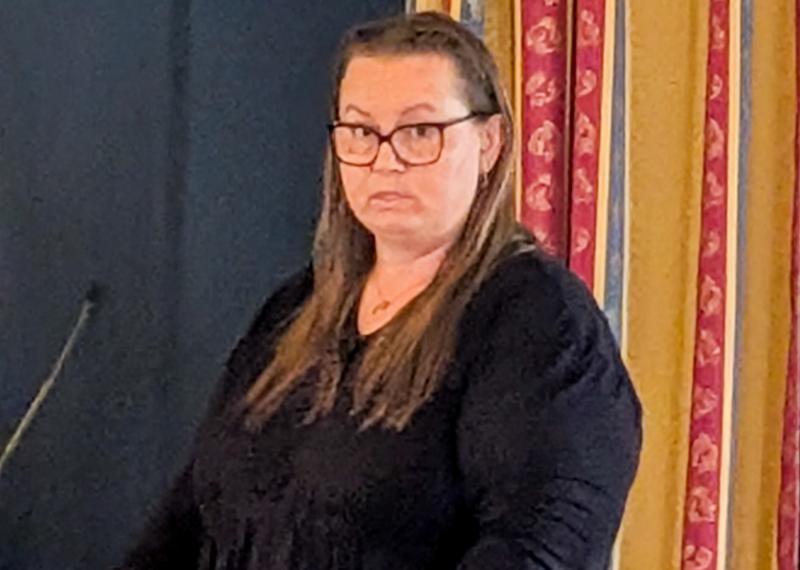 En kvinna med långt brunt hår och glasögon, klädd i en svart topp, står inomhus nära mönstrade gardiner och tittar mot kameran.