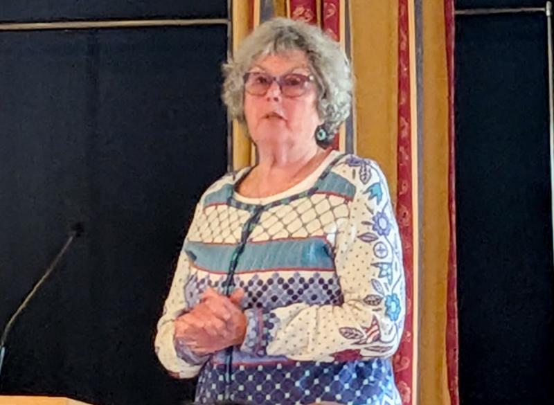 En äldre kvinna med grått lockigt hår och glasögon står och talar vid ett podium, iklädd en mönstrad tröja, framför en mörk och guldfärgad bakgrund.