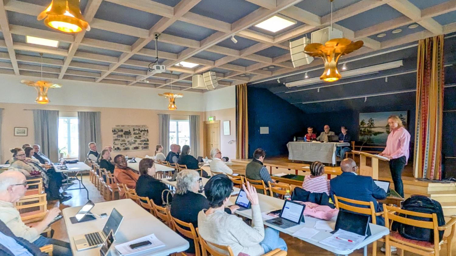 En grupp människor som sitter vid bord med bärbara datorer deltar i ett möte i en sal, medan en person står och talar vid ett podium nära en panel på fyra personer längst fram.