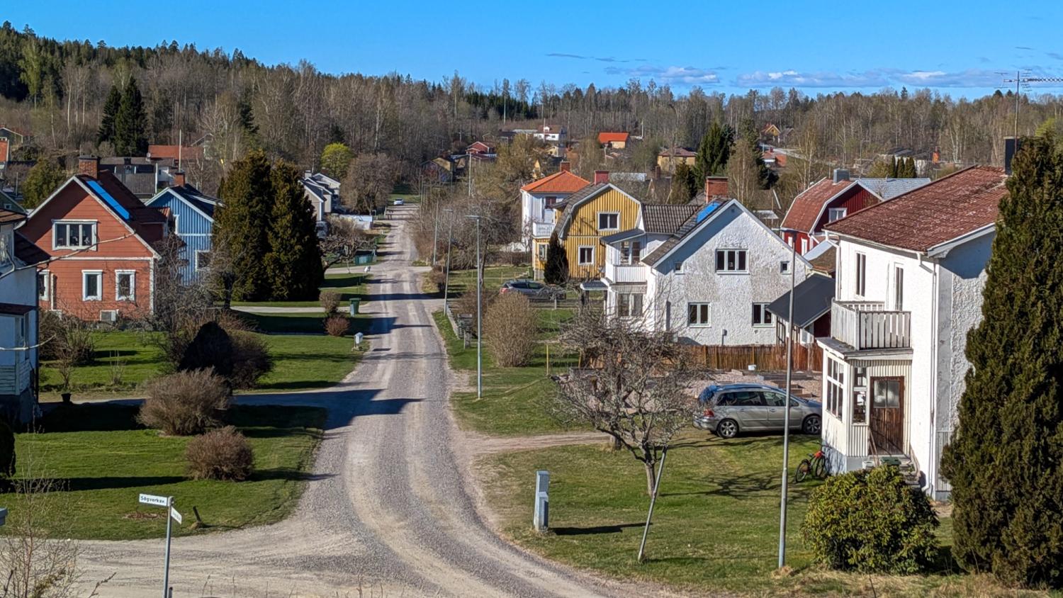 En lantlig bostadsgata med friliggande hus, gröna gräsmattor, träd och parkerade bilar under en klarblå himmel.