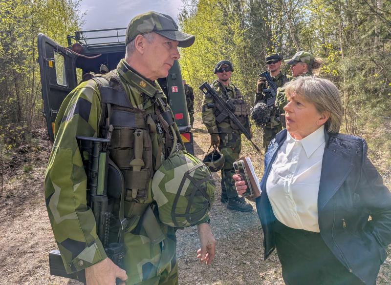 En kvinna intervjuar en uniformerad soldat som håller i en hjälm, medan andra beväpnade soldater står i närheten i en skogig utomhusmiljö.
