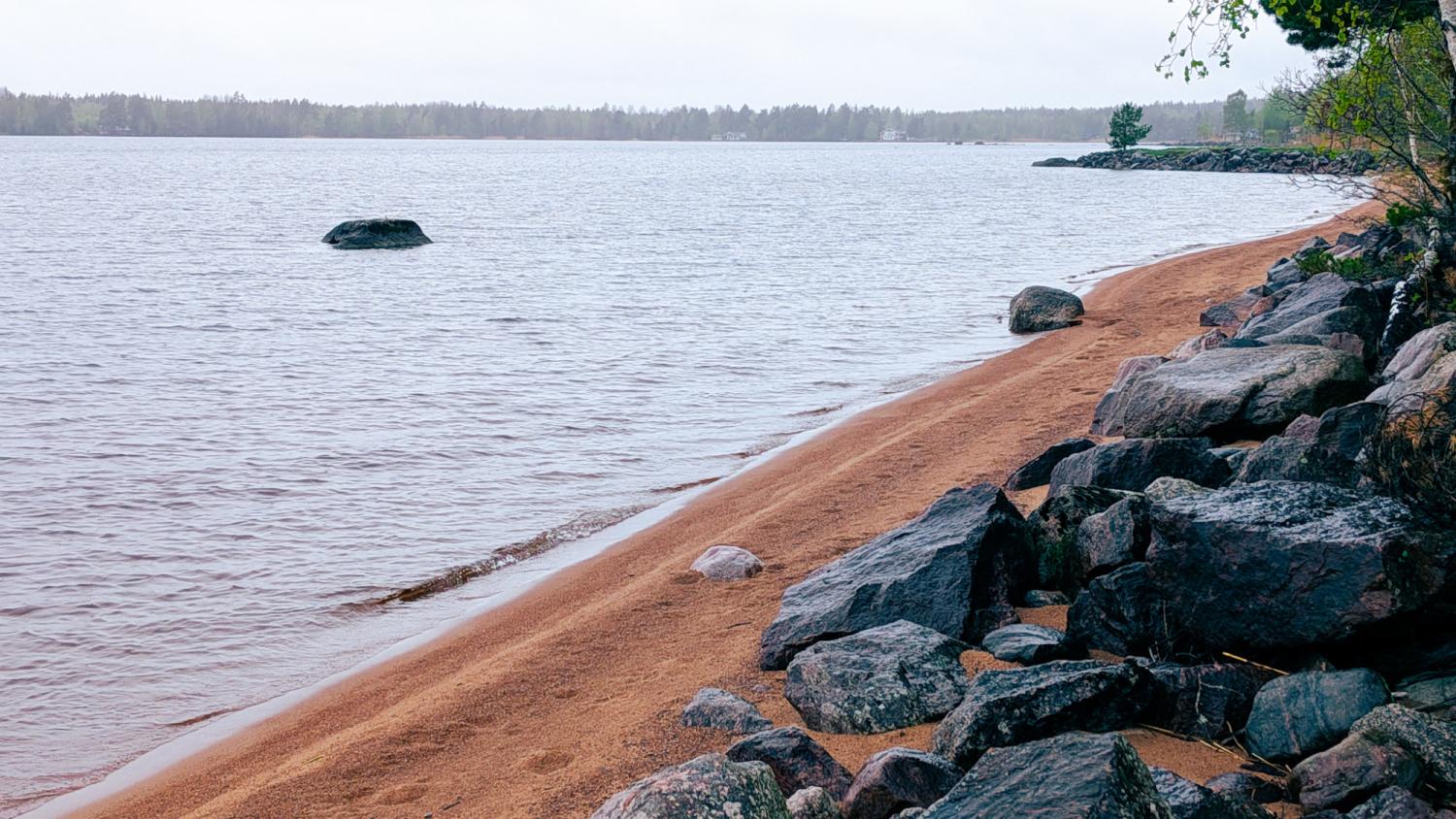 Klippig strandlinje med rödaktig sand vid ett lugnt vattendrag, utspridda stora stenar och träd i bakgrunden under en mulen himmel.