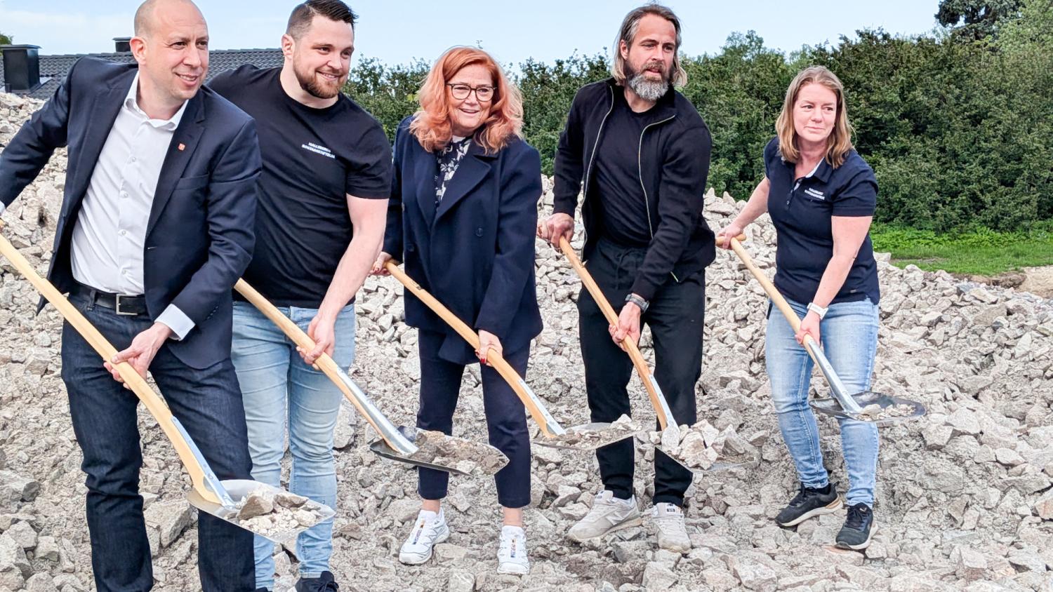 Fem personer står på en hög med stenar, var och en med en spade med stenar på, och ser ut att delta i ett första spadtag.