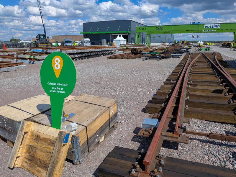 Järnvägsbygge med staplad räls, trälådor, ett krökt järnvägsspår och en grön skylt märkt 