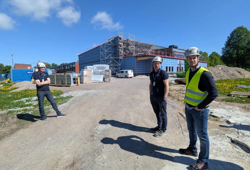 Tre män i skyddsutrustning står på en byggarbetsplats med en delvis uppförd byggnadsställning och byggmaterial i bakgrunden under en klarblå himmel.
