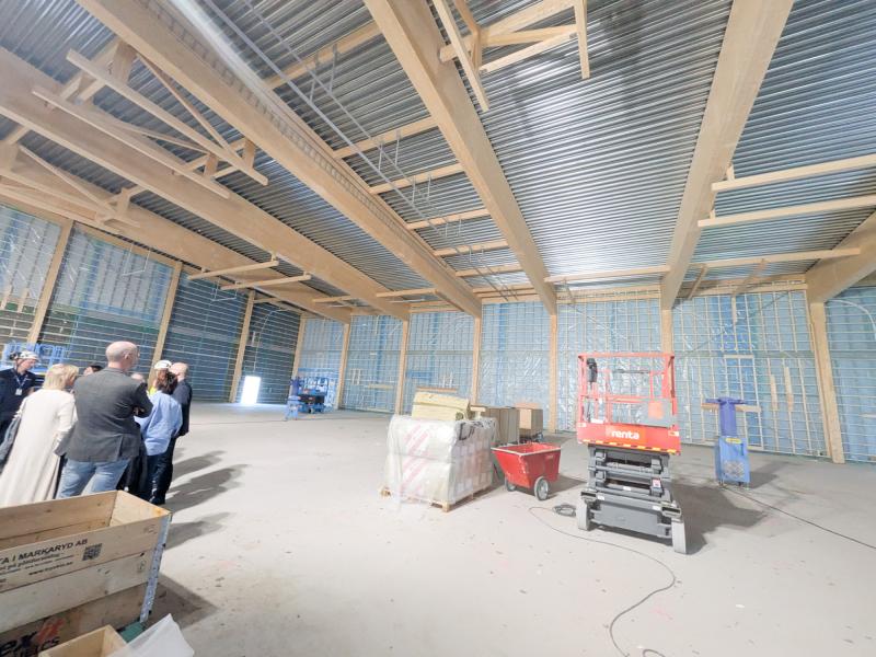 En grupp människor står inne i en stor, ofärdig byggnad med synliga bjälkar, byggmaterial och en röd saxlift.